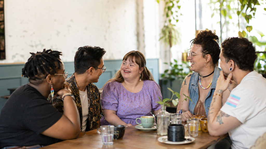 Diverse group of LGBTQ+ adults, including a woman with Down syndrome, sharing conversation and laughter at a café. Diverse group of LGBTQ+ adults, including a woman with Down syndrome, sharing conversation and laughter at a café.
